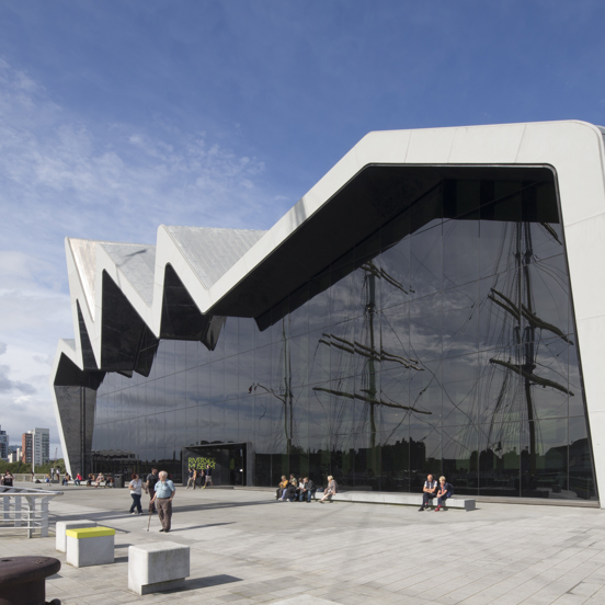 Sunny view of the modern, glass-fronted Riverside Museum, where the roofline reminiscent of a heartbeat on a monitor, with people enjoying the sunshine. The reflection of tall ship masts can be seen in the glass front of the museum